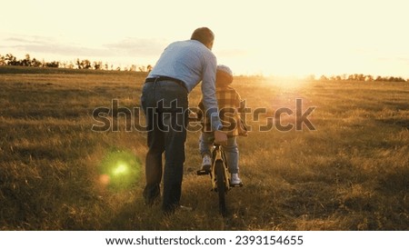Supportive father gives gentle push to help persistent son steer bicycle. Dad minimizes risk of falling holding bike with child during riding. Active kid enjoys fresh air with relative in nature Royalty-Free Stock Photo #2393154655