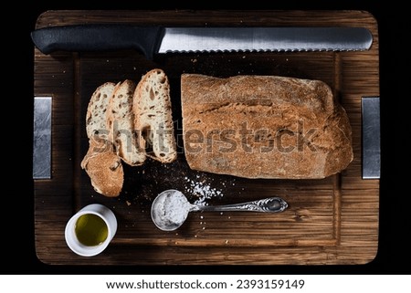 Composition with bread, knife, olive oil and coarse salt on wooden cutting board, top view