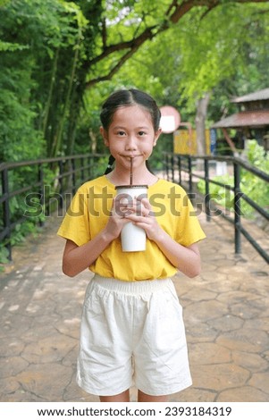 Portrait of happy Asian young girl kid holding paper cup and drinking cold water by straw with looking camera while standing in public garden.