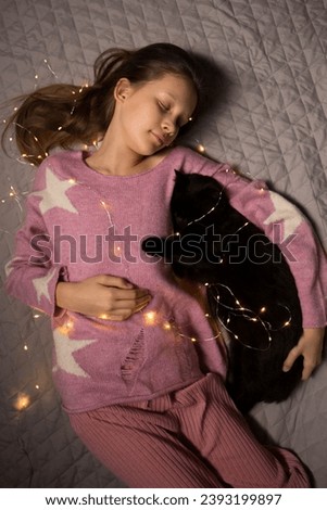 A girl in pink clothes and with a garland and black cat lies on the bed and dreams of Christmas