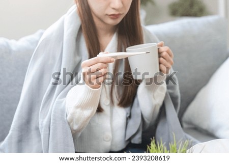 Sick, influenza asian young woman, girl have a fever, hand holding thermometer for check measure body temperature and drink hot water, illness while sitting rest on sofa at home. Health care concept. Royalty-Free Stock Photo #2393208619