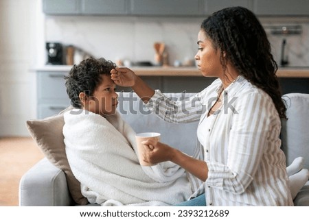 Caring Black Mother Giving Hot Tea To Her Ill Little Son And Touching His Forehead, Loving African American Mom Taking Care About Sick Male Child Suffering Seasonal Flu Or Cold, Closeup Royalty-Free Stock Photo #2393212689