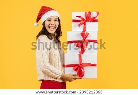 Happy young lady with a stack of Christmas gifts, beaming with smile in Santa hat expressing festive spirit, studio shot against yellow background, for holiday sale promotion