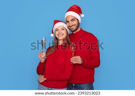 Happy Young Couple Wearing Santa Holding Champagne Glasses And Smiling At Camera, Cheerful Man And Woman Celebrating Christmas Holidays Together, Standing Isolated On Blue Background, Copy Space