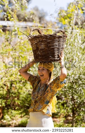 Farmer woman looking at the camera while carrying a wicker basket on her head outdoors in the garden.