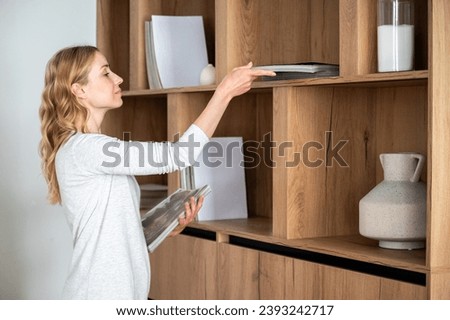 Young woman putting magazines and journals on wooden shelf of designer cabinet. Stylish bookcase with decorative elements in home library in living room. Leisure time.