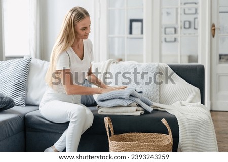 routine, house work, housekeeping concept. female housewife sitting on sofa at living room with cozy interior and folding her clothes, fresh wool sweater in stack after laundry Royalty-Free Stock Photo #2393243259