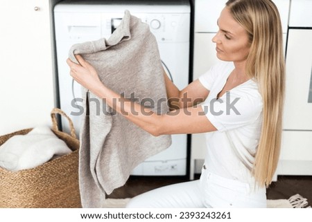 female housewife using detergent to remove stains, checking fresh soft wool sweater after washing in washer machine, sitting on floor near full wicker basket in room at modern apartment Royalty-Free Stock Photo #2393243261