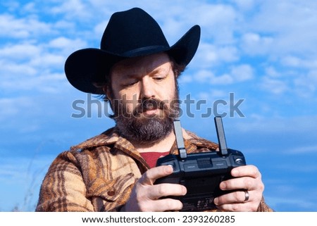 A modern cowboy wearing a black cowboy hat and a brown flannel jacket flies a drone with a modern controller as he looks at the screen in this closeup portrait with a blue sky on a sunny day.