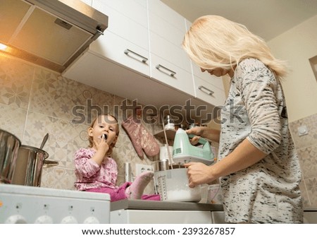 Little girl sitting on kitchen counter and helps her mommy make cookies  Royalty-Free Stock Photo #2393267857