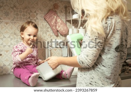 Little girl sitting on kitchen counter and helps her mommy make cookies  Royalty-Free Stock Photo #2393267859