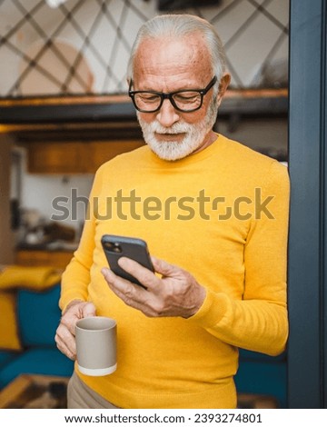 One senior man with a beard stand on the balcony hold a mobile phone and cup of coffee daily morning routine real person copy space read or use smartphone internet browse social network