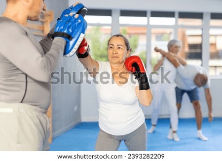 Active aged lady wearing boxing gloves practicing punches with trainer during sports classes