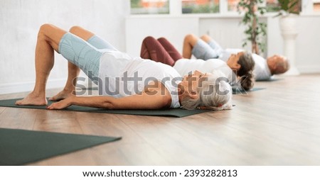 There is group Pilates class in studio for people of different ages. Elderly woman in company of hatha yoga lovers performs bridge pose, setu bandha sarvangasana Royalty-Free Stock Photo #2393282813