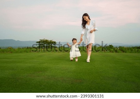 happy infant baby girl holding mother's hand and walking on a grass field with sky and mountain background