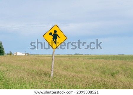 A caution school sign, at the side of the road, and a buildin far behind it on a field.