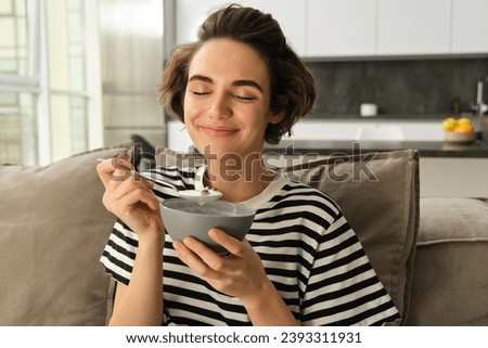 Portrait of young woman eating granola, bowl of cereals with milk, sitting on sofa and having her breakfast. Royalty-Free Stock Photo #2393311931