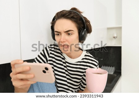 Portrait of woman with serious face, looking at smartphone with tensed emotion, watching interesting video on mobile phone, drinks tea, frowns and bites lips. Royalty-Free Stock Photo #2393311943
