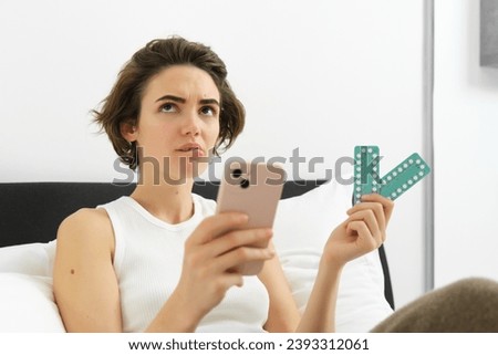 Portrait of thinking woman, lying in bed with pills, holding medication and mobile phone, searching for medicine dosage, feeling unwell at home, self-quarantine during covid-19. Royalty-Free Stock Photo #2393312061