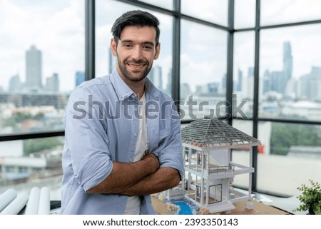 Portrait of architect engineer in casual outfit smile at camera while crossing arms. Businessman looking at camera and standing with arms folded near house model, architectural model. Tracery.