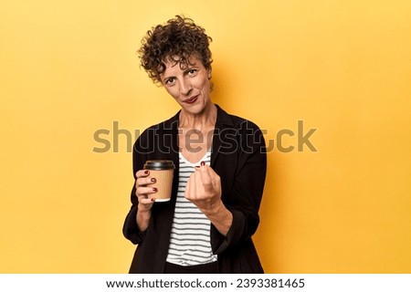 Businesswoman holding takeaway coffee on yellow pointing with finger at you as if inviting come closer.