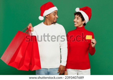 Merry young couple man woman wearing red casual clothes Santa hat posing hold shopping package bags isolated on plain green background. Black Friday sale buy day, Happy New Year 2024 Christmas concept