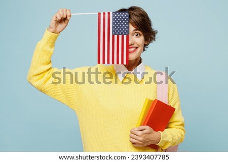 Young fun woman student wear casual clothes yellow sweater backpack bag hold cover half of face with notebook American flag isolated on plain blue background. High school university college concept