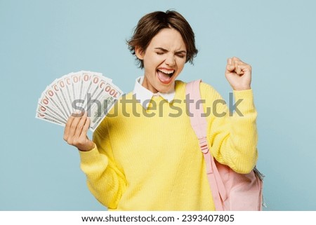 Young woman student wear casual clothes yellow sweater backpack bag hold fan cash money in dollar banknotes do winner gesture isolated on plain blue background. High school university college concept Royalty-Free Stock Photo #2393407805