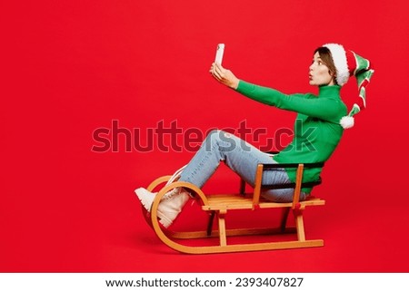 Full body young woman wears green turtleneck Santa hat posing sit ride sled do selfie shot mobile cell phone isolated on plain red background. Happy New Year 2024 celebration Christmas holiday concept