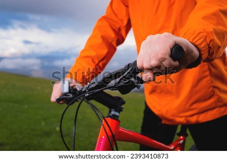 Close up, hands of a cyclist on the handlebars of a mountain bike on a green natural background and cloudy sky. Taking a break while riding a dirt bike