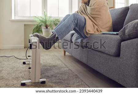 Side view of a frozen man sitting on sofa in the living room at home wrapped in blanket and trying to warm his feet in warm socks putting his legs on electric heater. Heating problems concept.