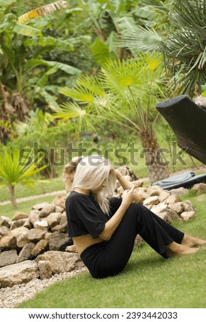 Blond young woman sitting outside on grass looking at phone side profile palm trees in background in Thailand barefoot holding one hand on her head