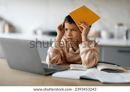 Learning Difficulties. Stressed Little Black Boy Using Laptop And Covering Head With Workbook At Home, Annoyed Preteen Male Child Sitting At Desk With Computer, Suffering Problems With Homework Royalty-Free Stock Photo #2393453243
