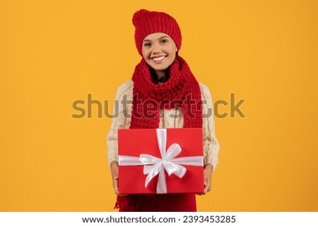 Positive young woman in red knitwear holding wrapped Christmas gift box, smiling to camera offering present. Banner for Xmas And New Year celebrations, studio shot against yellow background