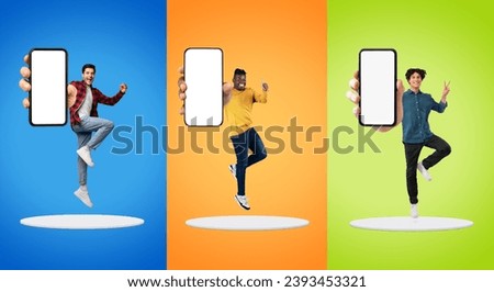 Cheerful young diverse men show big smartphone with blank screen, jump at platform, have fun, peace, make thumb up, victory sign, isolated on multicolored background. Win, app recommendation