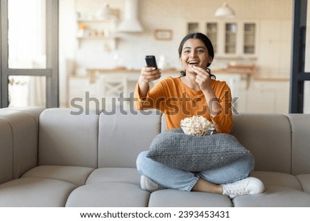 Happy Young Indian Woman Holding Remote Controller And Eating Popcorn At Home, Cheerful Millennial Eastern Lady Watching TV Show While Relaxing On Couch In Cozy Living Room, Copy Space Royalty-Free Stock Photo #2393453431