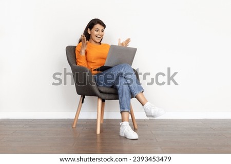 Emotional millennial indian lady sitting in armchair over white blank wall background, looking at laptop screen, screaming and gesturing, reading great news, got good job opportunity, copy space