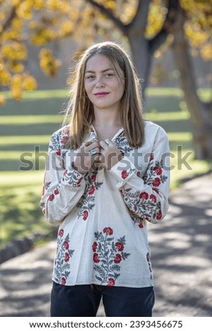 Beneath the autumn trees, a young woman buttons up her shirt as she looks to at the camera
