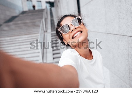 Young beautiful smiling hipster woman in trendy summer white t-shirt and jeans clothes. Carefree woman, posing in the street at sunny day. Positive model outdoors. Cheerful and happy. Takes pov selfie