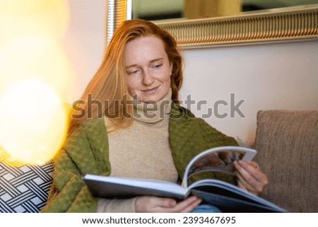 Ginger red haired woman watching family pictures from photo book. Red-headed cosy mother wrapped with blanket at home on sofa memories during Christmas holiday. New year cozy recreation in winter