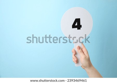 Woman holding auction paddle with number 4 on light blue background, closeup. Space for text Royalty-Free Stock Photo #2393469461