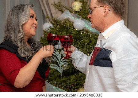 Waist up portrait of romantic scene of elegant couple toasting with red wine glasses welcoming new year, Christmas tree with blurred background, formal clothes, happy smiling expression