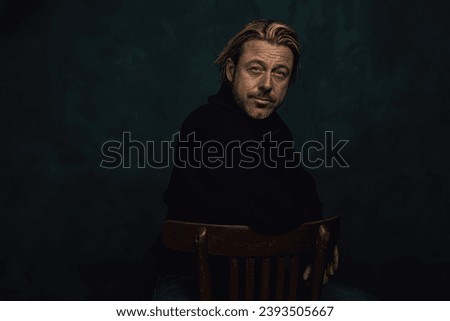 Middle aged blond caucasian man with stubble beard in black woolen jersey sits on wooden chair in front of a dark wall.