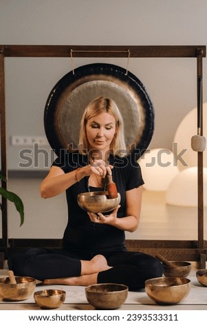 A woman plays a Tibetan singing bowl while sitting on a yoga mat against the background of a gong.
