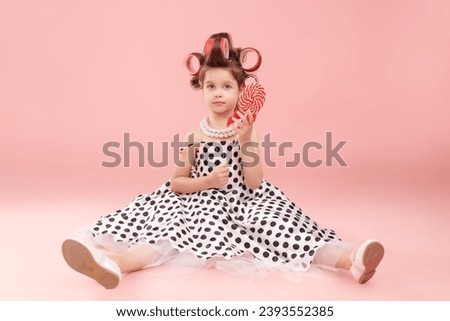 Little fashionista girl dresses up for a party and holds a lollipop. Pink studio background. Kid's fashion. Pin-up style. Sweets for children.