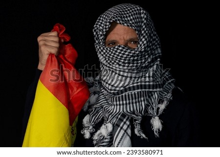 Adult man with Palestinian scarf put on his head on a black background holding a flag of Spain Royalty-Free Stock Photo #2393580971
