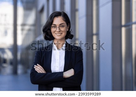 Close-up portrait of a young businesswoman standing outside an office building with her arms crossed over her chest. Confidently and smilingly looking at the camera. Royalty-Free Stock Photo #2393599289