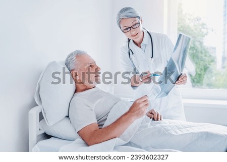Adult doctor standing in ward near elderly patient showing picture of an x-ray. Bones and lungs image. Doctor explaining telling the diagnosis to grandfather