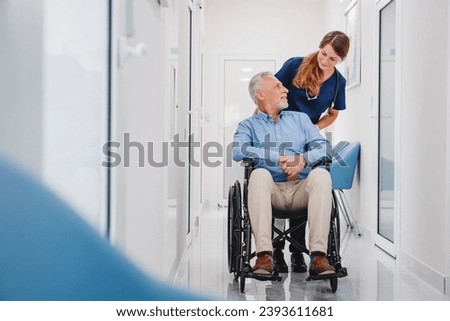 Senior male patient sitting in wheelchair in hospital corridor with female nurse doctor. Caregiver medical worker supporting disabled immobile patient in hospice Royalty-Free Stock Photo #2393611681