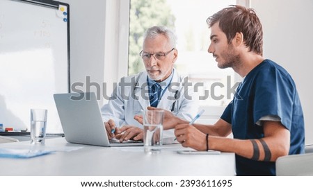 Two healthcare workers in consultation room using laptop and discussing work. Medical student studying, having intern practice in hospital, talking about diagnosis
